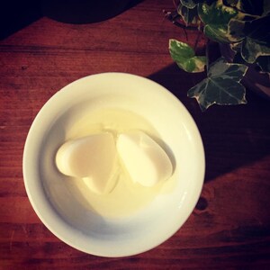 May include: Two white heart-shaped wax melts in a white ceramic bowl with a yellow liquid. The bowl is on a wooden surface.