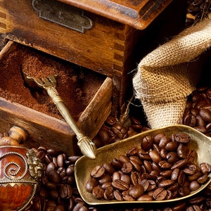 May include: A vintage wooden coffee grinder with a drawer full of ground coffee beans. A brass scoop filled with whole coffee beans sits on a bed of coffee beans. A burlap sack is in the background.