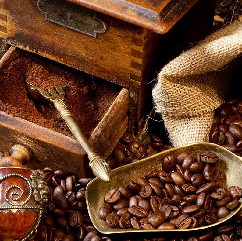 May include: A vintage wooden coffee grinder with a drawer full of ground coffee beans. A brass scoop filled with whole coffee beans sits on a bed of coffee beans. A burlap sack is in the background.