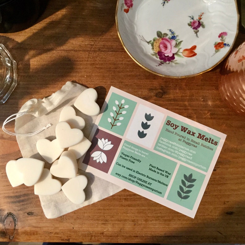 May include: A white muslin bag filled with heart-shaped soy wax melts. The bag is sitting on a wooden surface next to a card that reads "Soy Wax Melts" and includes information about the product, including that it is hand-poured in small batches, vegan friendly, plant-based wax, plastic free, made in the UK, and can be used in electric burners/melters. The card also includes the company's website and social media handles.
