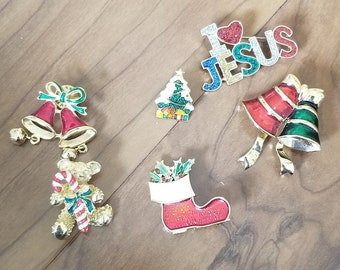 May include: A collection of six Christmas-themed pins, including a stocking, a teddy bear holding a candy cane, two bells, a Christmas tree, and a pin that says "I Love Jesus".
