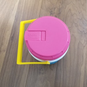 May include: A pink Igloo water cooler with a yellow handle. The cooler has a white base and a blue stripe around the middle. The lid has a latch with the text "Igloo 1/2 Gallon" on it.