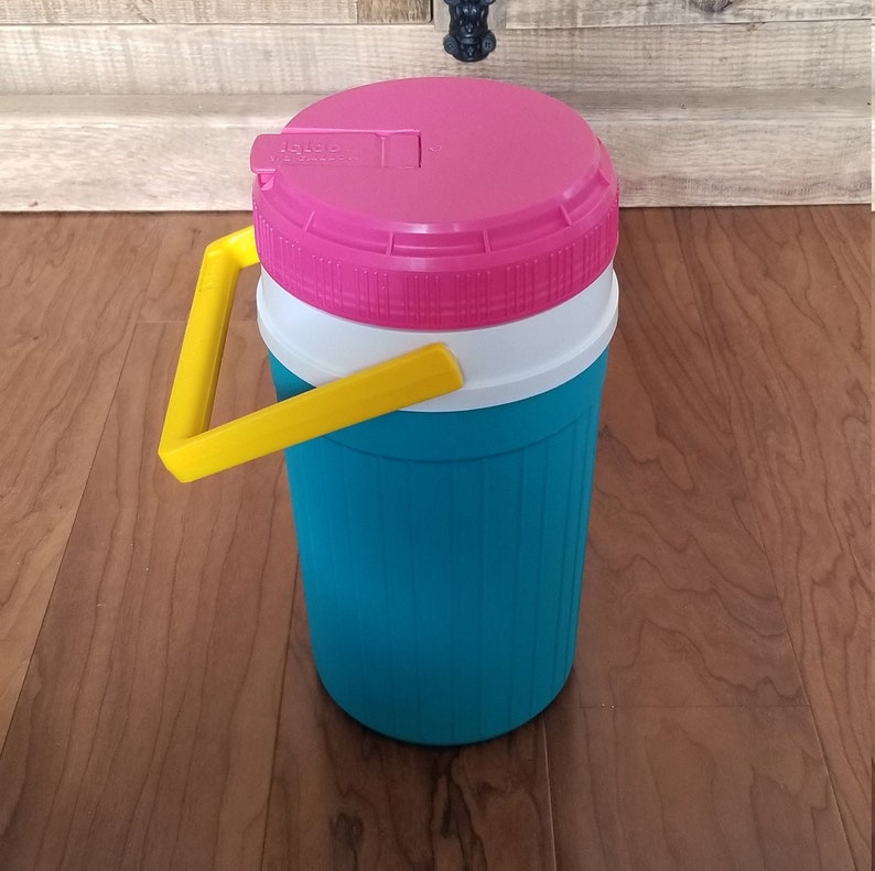 May include: A teal and white insulated beverage container with a pink lid and a yellow handle. The lid has the word "IGLOO" printed on it.