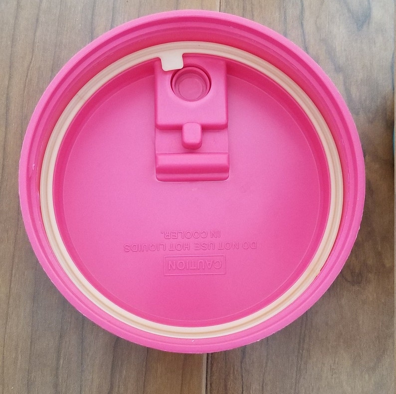 May include: Pink lid for a cooler with a white seal and a latch. The lid has the text "CAUTION DO NOT USE HOT LIQUIDS IN COOLER".