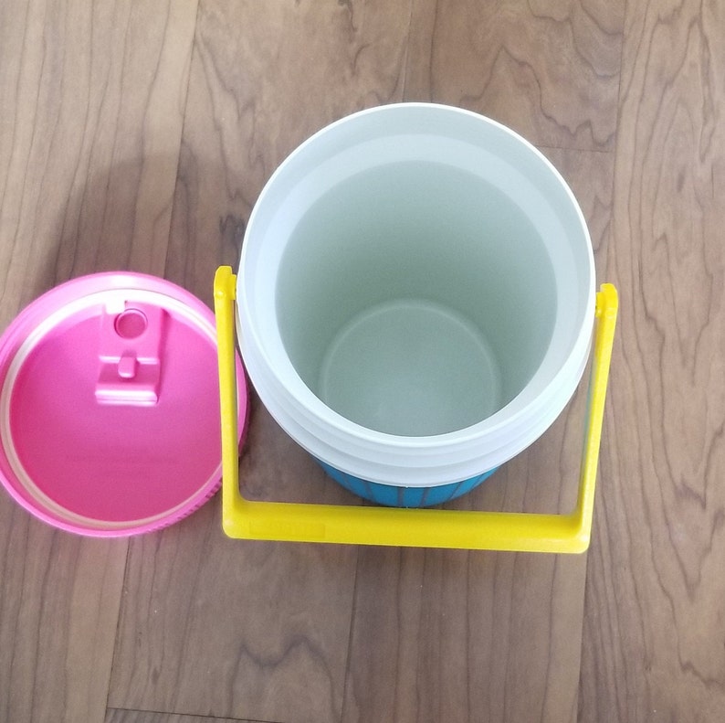 May include: A blue and yellow insulated drinks container with a white lid and a pink lidded cup. The container has a yellow handle.
