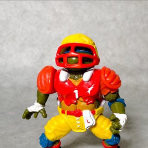 K&ouml;nnte beinhalten: Eine Teenage Mutant Ninja Turtles Actionfigur, die eine rot-gelb-blaue Footballuniform mit Helm tr&auml;gt. Die Figur steht mit ausgestreckten Armen.
