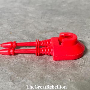 Puede incluir: Una nave espacial de juguete de plástico rojo con dos cañones en la parte delantera. La nave espacial tiene una forma redondeada y una pequeña abertura en la parte superior.