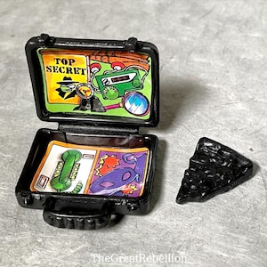 May include: A black plastic briefcase with a handle, open to reveal a cartoon image of a spy with the words "Top Secret". The briefcase has a green and yellow interior with a cartoon image of a phone, a magnifying glass, and a radio.