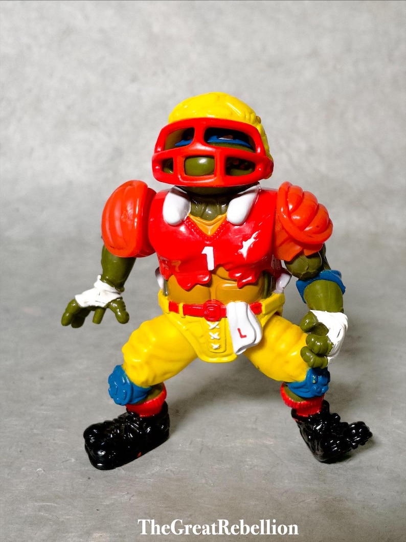 K&ouml;nnte beinhalten: Eine Teenage Mutant Ninja Turtles Actionfigur, die eine rot-gelb-blaue Footballuniform mit Helm tr&auml;gt. Die Figur steht mit ausgestreckten Armen.