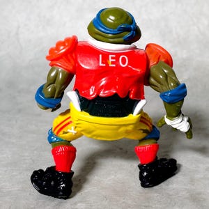 K&ouml;nnte beinhalten: Eine gr&uuml;ne, rote, gelbe und blaue Teenage Mutant Ninja Turtles Actionfigur, die ein Football-Uniform mit dem Namen "LEO" auf der Vorderseite tr&auml;gt.