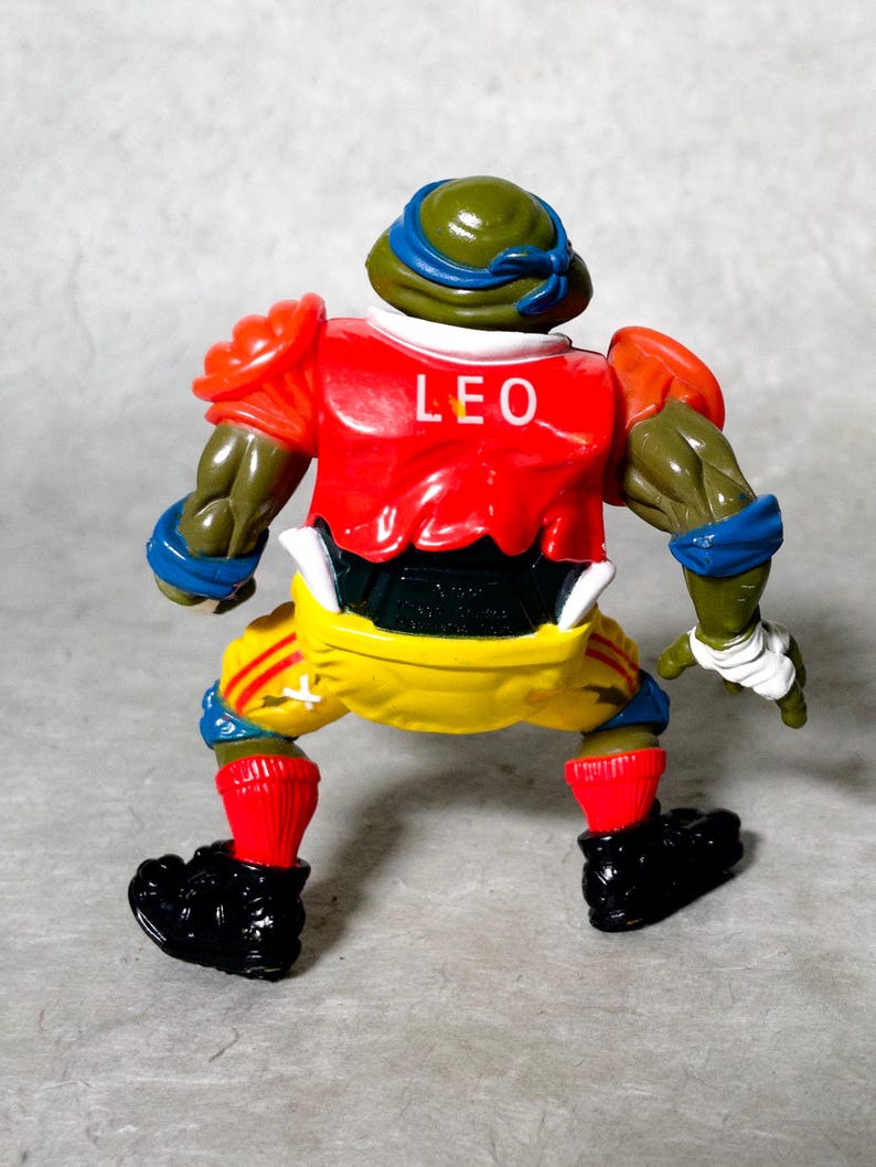 K&ouml;nnte beinhalten: Eine gr&uuml;ne, rote, gelbe und blaue Teenage Mutant Ninja Turtles Actionfigur, die ein Football-Uniform mit dem Namen "LEO" auf der Vorderseite tr&auml;gt.
