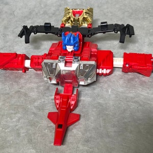 Puede incluir: Un juguete robot rojo y plateado con una cabeza azul y alas negras. El robot est&aacute; en modo robot con los brazos y las piernas extendidos. El robot tiene una placa de pecho plateada con un dise&ntilde;o rojo y plateado.