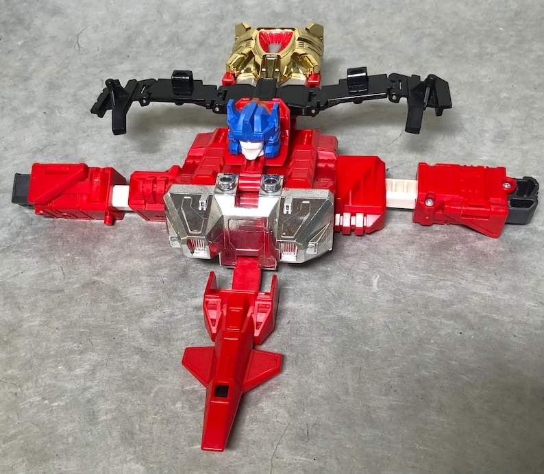 Puede incluir: Un juguete robot rojo y plateado con una cabeza azul y alas negras. El robot est&aacute; en modo robot con los brazos y las piernas extendidos. El robot tiene una placa de pecho plateada con un dise&ntilde;o rojo y plateado.