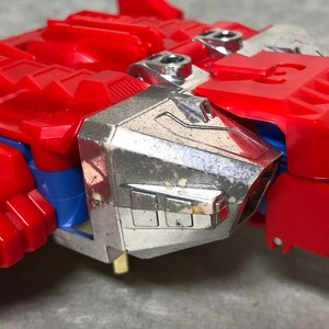 Puede incluir: Un robot de juguete rojo y plateado con un dise&ntilde;o detallado. El robot tiene una cabeza plateada y un cuerpo rojo con una franja azul. El robot est&aacute; en modo robot.