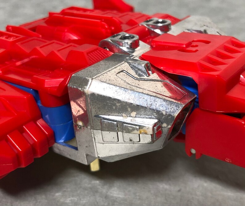 Puede incluir: Un robot de juguete rojo y plateado con un dise&ntilde;o detallado. El robot tiene una cabeza plateada y un cuerpo rojo con una franja azul. El robot est&aacute; en modo robot.