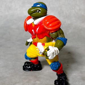 K&ouml;nnte beinhalten: Eine gr&uuml;ne Teenage Mutant Ninja Turtles Actionfigur, die ein rot-gelb-blaues Football-Uniform tr&auml;gt. Die Figur ist in einer Laufhaltung mit einem Bein nach vorne und dem anderen Bein nach hinten aufgestellt. Die Figur hat einen gr&uuml;nen Kopf mit einem blauen Bandana und einer wei&szlig;en Gesichtsmaske.