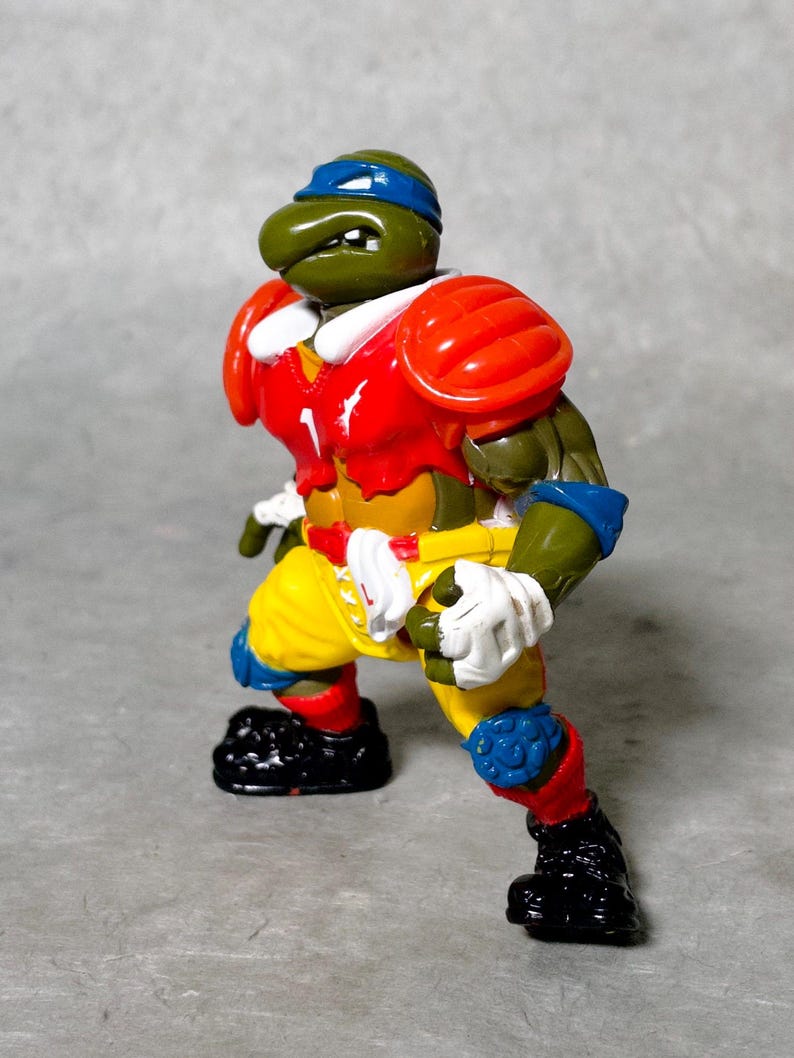 K&ouml;nnte beinhalten: Eine gr&uuml;ne Teenage Mutant Ninja Turtles Actionfigur, die ein rot-gelb-blaues Football-Uniform tr&auml;gt. Die Figur ist in einer Laufhaltung mit einem Bein nach vorne und dem anderen Bein nach hinten aufgestellt. Die Figur hat einen gr&uuml;nen Kopf mit einem blauen Bandana und einer wei&szlig;en Gesichtsmaske.