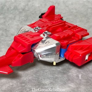 Puede incluir: Un robot de juguete rojo y plateado en su modo veh&iacute;culo. El robot tiene la forma de un avi&oacute;n a reacci&oacute;n futurista con una nariz puntiaguda y alas. El robot tiene un pecho plateado y un cuerpo rojo.