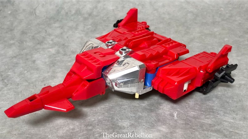 Puede incluir: Un robot de juguete rojo y plateado en su modo veh&iacute;culo. El robot tiene la forma de un avi&oacute;n a reacci&oacute;n futurista con una nariz puntiaguda y alas. El robot tiene un pecho plateado y un cuerpo rojo.