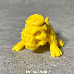 May include: A yellow plastic toy bulldog with a yellow cap. The bulldog has a fierce expression and is in a crouching position.