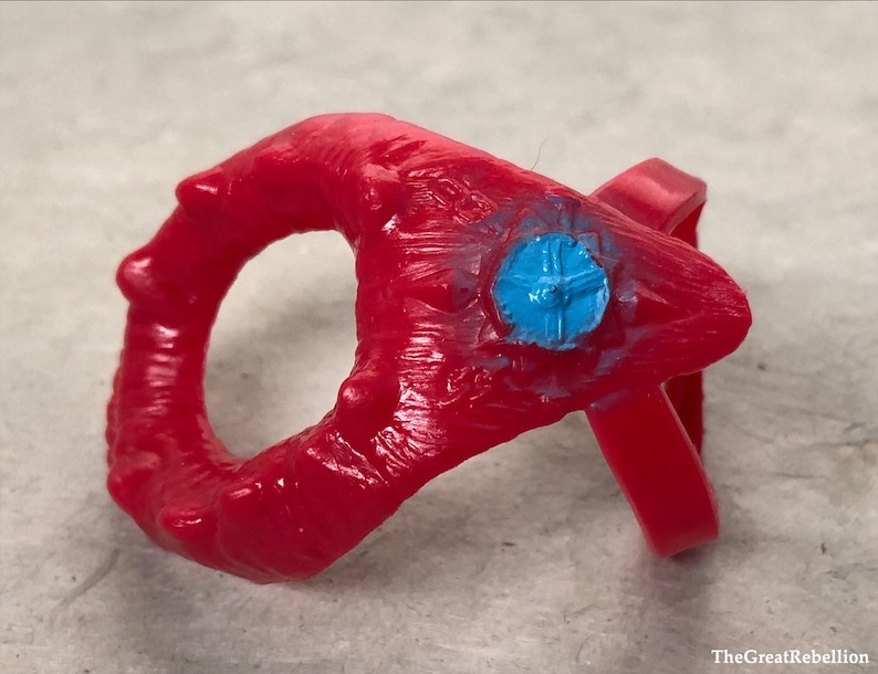 May include: A red plastic ring with a blue plastic centre piece. The ring has a textured surface and is decorated with small spikes.