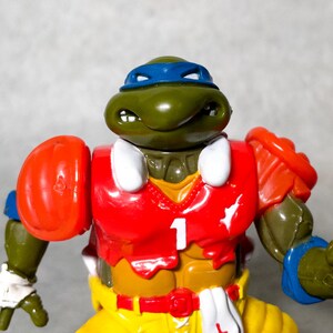 K&ouml;nnte beinhalten: Gr&uuml;ne Teenage Mutant Ninja Turtles Actionfigur, die ein rot-wei&szlig;es Football-Trikot mit der Nummer "1" auf der Vorderseite tr&auml;gt. Die Figur hat ein blaues Bandana und h&auml;lt einen Football.