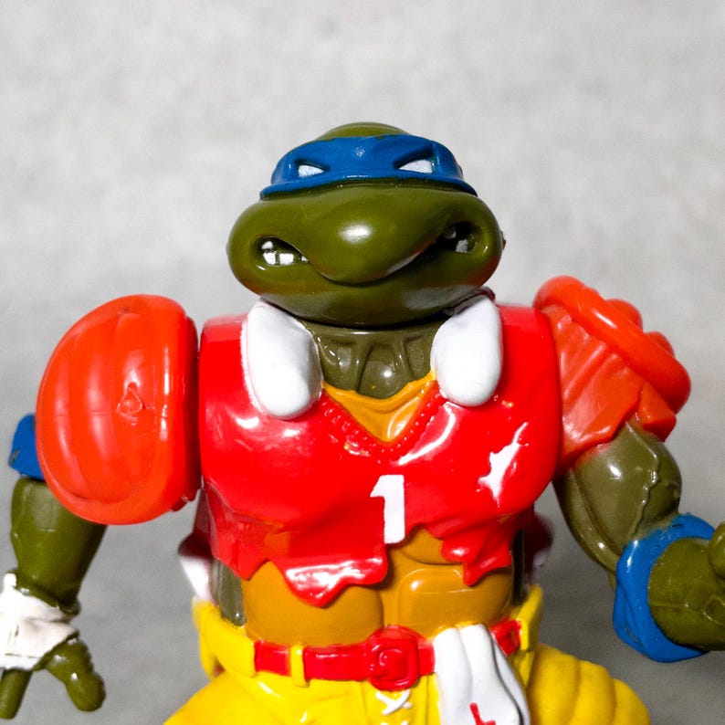 K&ouml;nnte beinhalten: Gr&uuml;ne Teenage Mutant Ninja Turtles Actionfigur, die ein rot-wei&szlig;es Football-Trikot mit der Nummer "1" auf der Vorderseite tr&auml;gt. Die Figur hat ein blaues Bandana und h&auml;lt einen Football.