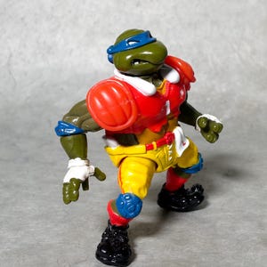 K&ouml;nnte beinhalten: Eine gr&uuml;ne Teenage Mutant Ninja Turtles Actionfigur, die ein rot-wei&szlig;-blaues Football-Uniform tr&auml;gt. Die Figur hat eine blaue Maske und h&auml;lt einen Football.
