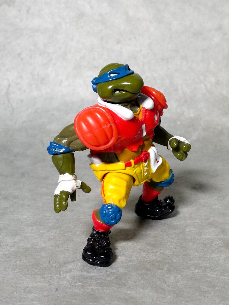 K&ouml;nnte beinhalten: Eine gr&uuml;ne Teenage Mutant Ninja Turtles Actionfigur, die ein rot-wei&szlig;-blaues Football-Uniform tr&auml;gt. Die Figur hat eine blaue Maske und h&auml;lt einen Football.