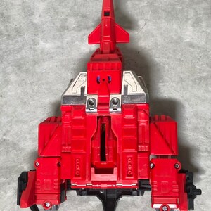 Puede incluir: Un robot de juguete rojo y plateado en su forma de nave espacial. El robot est&aacute; en medio de transformarse en su forma de robot. La nave espacial tiene una nariz puntiaguda y alas. El robot tiene un pecho grande y brazos.