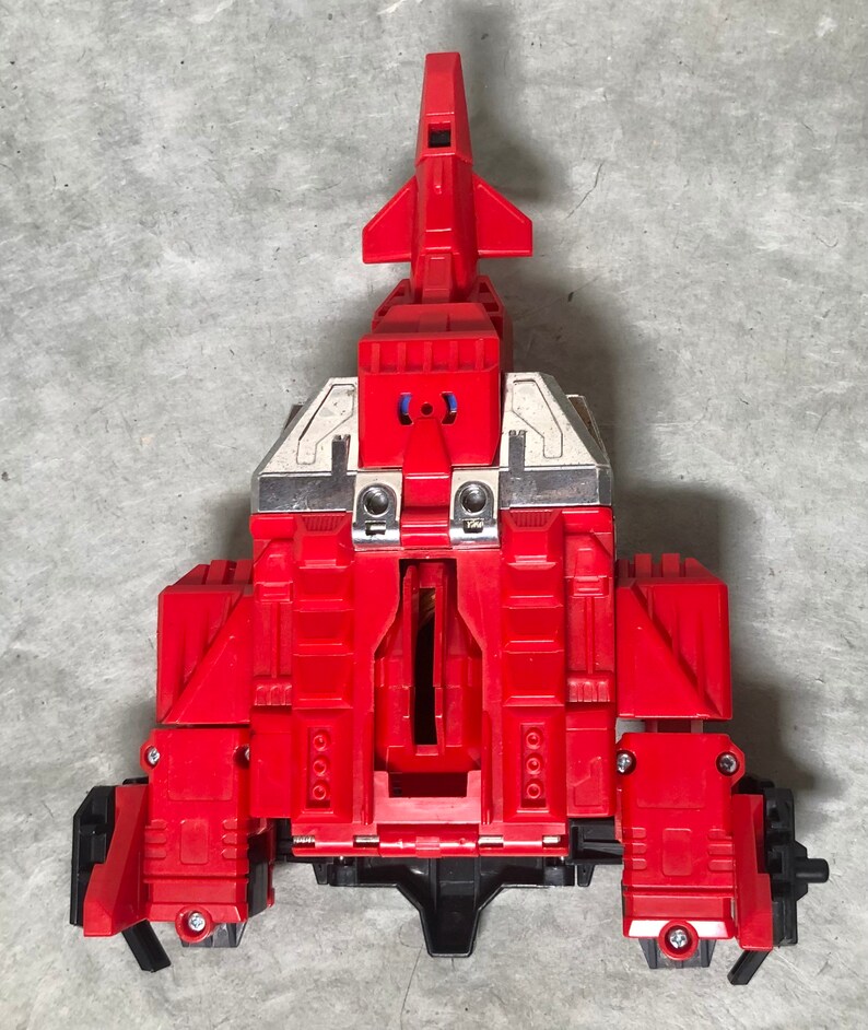 Puede incluir: Un robot de juguete rojo y plateado en su forma de nave espacial. El robot est&aacute; en medio de transformarse en su forma de robot. La nave espacial tiene una nariz puntiaguda y alas. El robot tiene un pecho grande y brazos.