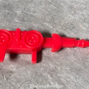 Puede incluir: Un arma de juguete de plástico rojo brillante con una hélice en el extremo. El juguete tiene un cuerpo rectangular con dos elementos circulares y dos patas pequeñas. Es un artículo vintage.