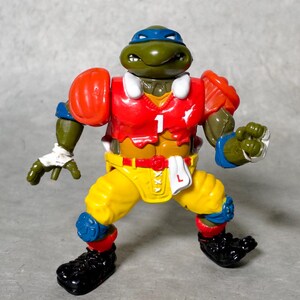 K&ouml;nnte beinhalten: Eine gr&uuml;ne Teenage Mutant Ninja Turtles Actionfigur, die ein rot-gelb-blaues Football-Trikot mit der Nummer "1" auf der Vorderseite tr&auml;gt. Die Figur hat ein gr&uuml;nes Gesicht mit einem blauen Bandana und h&auml;lt einen Football.