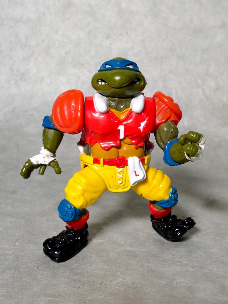 K&ouml;nnte beinhalten: Eine gr&uuml;ne Teenage Mutant Ninja Turtles Actionfigur, die ein rot-gelb-blaues Football-Trikot mit der Nummer "1" auf der Vorderseite tr&auml;gt. Die Figur hat ein gr&uuml;nes Gesicht mit einem blauen Bandana und h&auml;lt einen Football.
