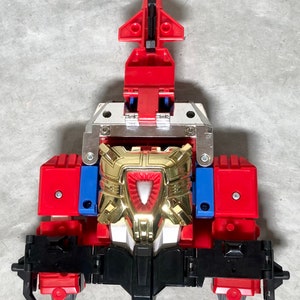 Puede incluir: Un juguete robot rojo, dorado y negro con una placa de pecho plateada. El robot tiene una cabeza puntiaguda y una base negra.