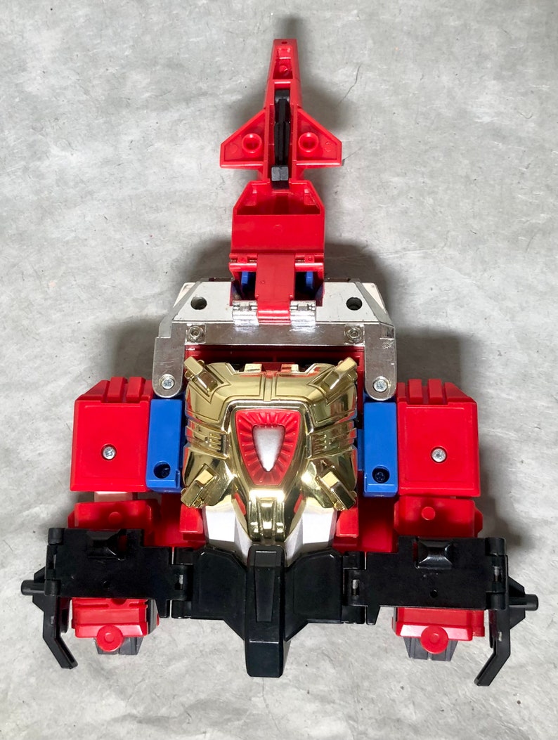 Puede incluir: Un juguete robot rojo, dorado y negro con una placa de pecho plateada. El robot tiene una cabeza puntiaguda y una base negra.