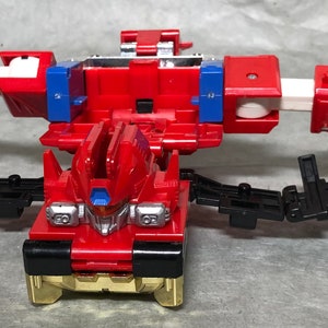 Puede incluir: Un robot de juguete rojo y negro con brazos y piernas articulados. El robot est&aacute; en un estado transformado, con los brazos extendidos hacia afuera y las piernas separadas. El robot tiene un pecho dorado y una cabeza plateada.