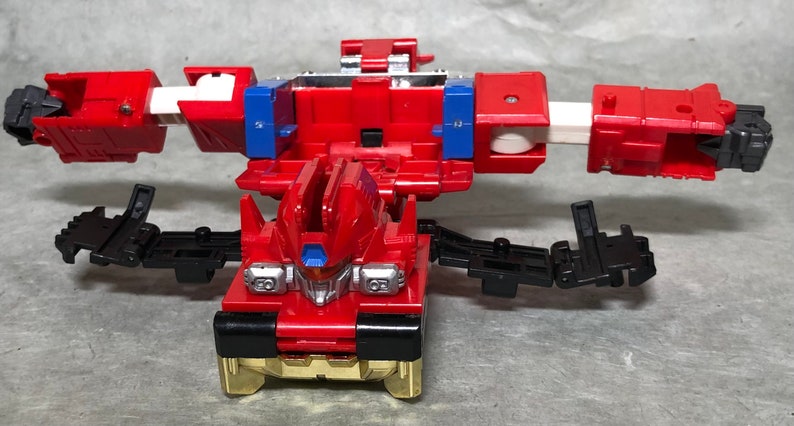 Puede incluir: Un robot de juguete rojo y negro con brazos y piernas articulados. El robot est&aacute; en un estado transformado, con los brazos extendidos hacia afuera y las piernas separadas. El robot tiene un pecho dorado y una cabeza plateada.