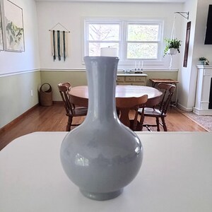 May include: A light blue ceramic vase with a rounded body and a long, slender neck. The vase is displayed on a white surface, with a blurred background of a dining room featuring a wooden table and chairs.