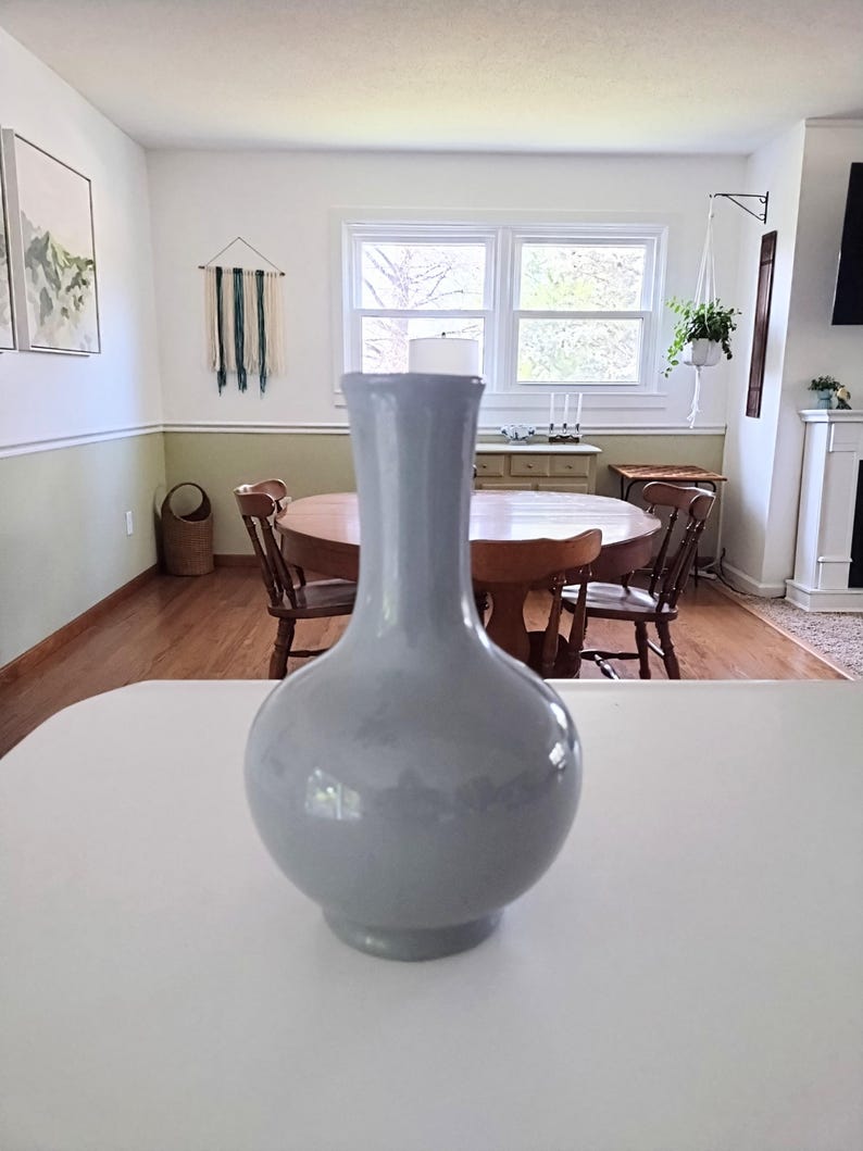 May include: A light blue ceramic vase with a rounded body and a long, slender neck. The vase is displayed on a white surface, with a blurred background of a dining room featuring a wooden table and chairs.