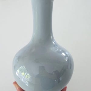 May include: A light blue ceramic vase with a bulbous body and a slender neck. The vase has a glossy finish and sits on a small, circular base. The vase is held by a person's hand.