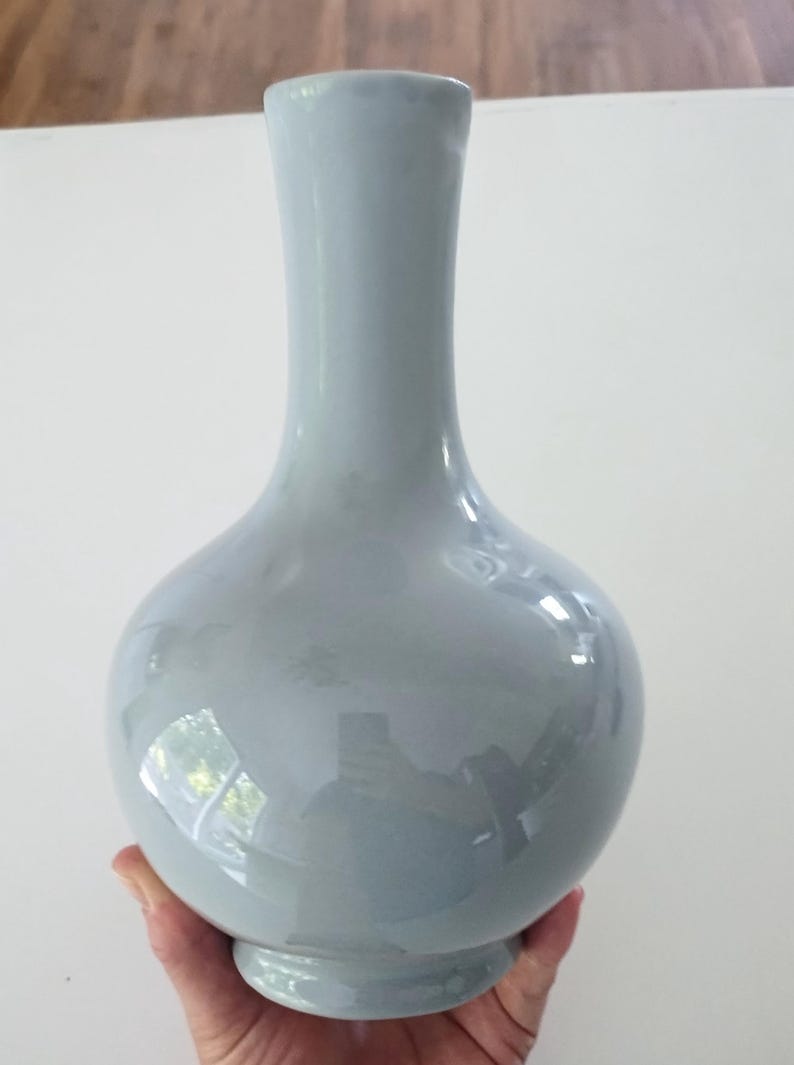 May include: A light blue ceramic vase with a bulbous body and a slender neck. The vase has a glossy finish and sits on a small, circular base. The vase is held by a person's hand.