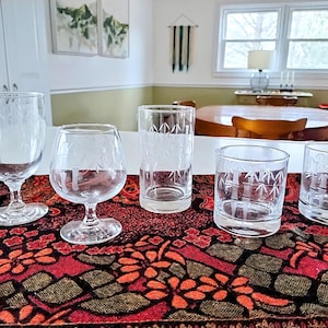 May include: Five clear glass drinking glasses with etched bamboo designs. The glasses are arranged on a red and black patterned table runner.