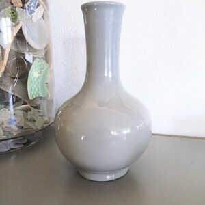 May include: A gray ceramic vase with a rounded body and a long, slender neck. The vase has a glossy finish and sits on a gray surface. The vase is a decorative item.