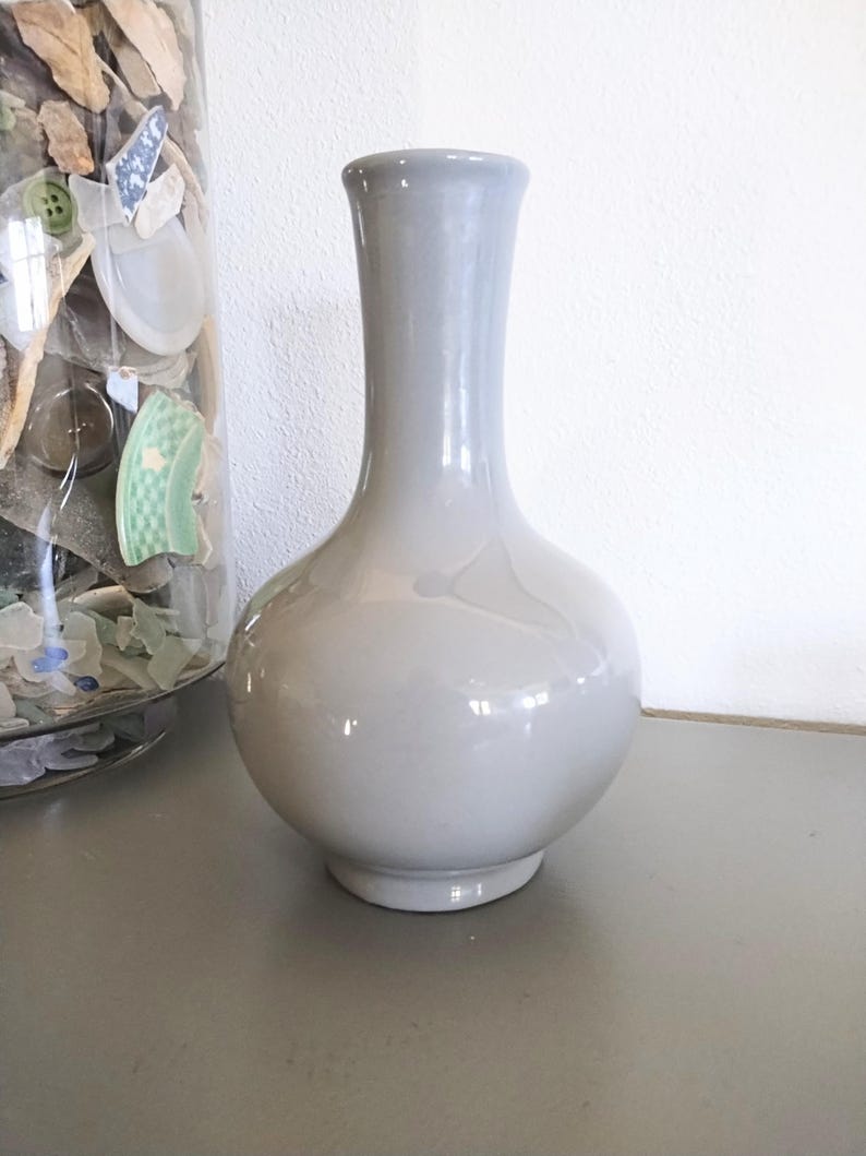 May include: A gray ceramic vase with a rounded body and a long, slender neck. The vase has a glossy finish and sits on a gray surface. The vase is a decorative item.