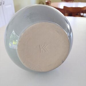 May include: A light blue ceramic vase with a round body and a flat, unglazed base. The base has a stamped "K" mark. The vase is sitting on a white surface.