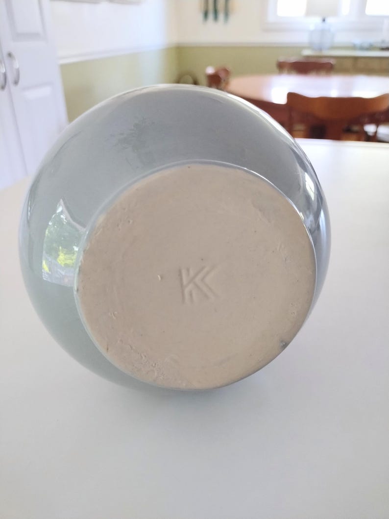 May include: A light blue ceramic vase with a round body and a flat, unglazed base. The base has a stamped "K" mark. The vase is sitting on a white surface.