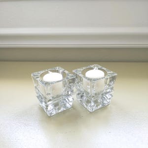 May include: Two clear, square glass candle holders with a stacked design. Each holder contains a white, round candle. The candle holders are on a light-colored surface, with a white wall in the background.