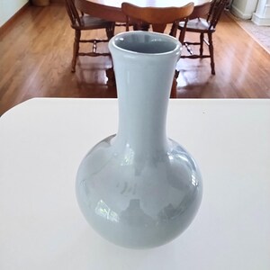 May include: A light blue ceramic vase with a rounded base and a slender neck. The vase is displayed on a white surface, with a wooden table and chairs visible in the background. The vase is approximately 10 inches tall.