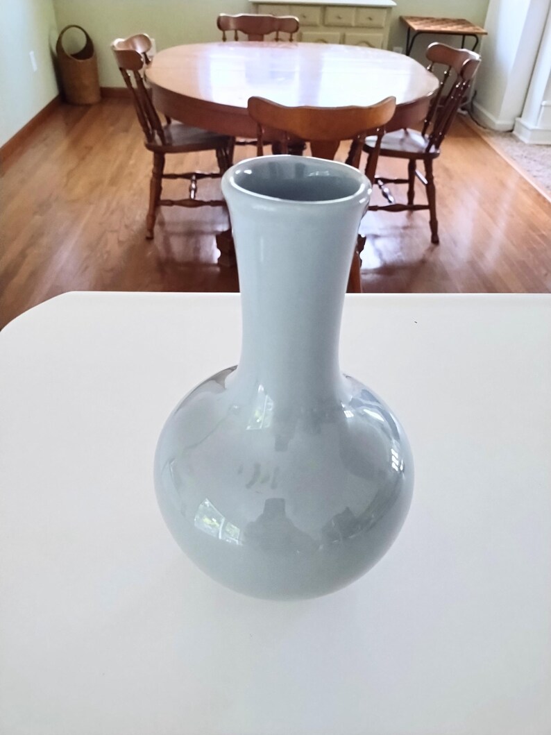 May include: A light blue ceramic vase with a rounded base and a slender neck. The vase is displayed on a white surface, with a wooden table and chairs visible in the background. The vase is approximately 10 inches tall.
